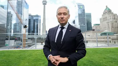 Valerie Macon/AFP via Getty Images Champagne wears a suit and tie while he stands on a lawn in front of the Toronto city skyline of high-rise buildings.