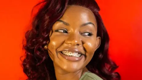 That Girl Boina A young woman in braces and a burgundy wig, wearing a green vest, smiling away from the camera with her head tilted to the left behind a red background