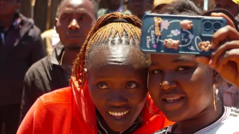 A close-up of two young women taking a selfie. One is wearing an orange top and the other a white T-shirt.