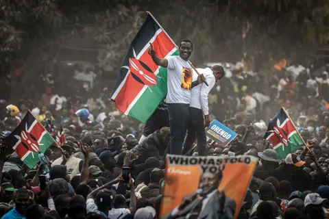 AFP via Getty Images Edwin Sifuna in a white T-shirt stands on top of a car and holds a Kenyan flag as crowds gather round. 