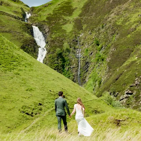 Jana Rukute Angelika is in her wedding dress and Patryk is in his green wedding suit as they stand gazing at a waterfall. 