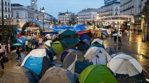 Getty Images About a dozen small tents are pitched in a concrete square in the centre of Madrid. It is raining and people walk around the tens with umbrellas. 