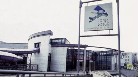 The entrance to Ponds Forge International Sports Centre in Sheffield is glass-fronted with white brick on either side. A purple water slide snakes out of the building and back in again, on the left-hand side of the image.