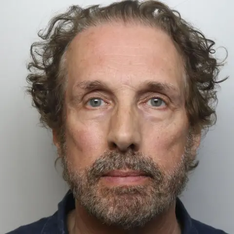 South Yorkshire Police A man with blue-grey eyes, a grey beard and curly brown hair is shown in a police custody image
