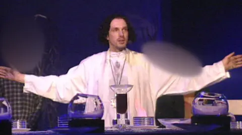 Nine O'Clock Trust Chris Brain leads a service, wearing white robes and with his arms out wide. The lighting is dim with a blue hue.