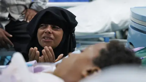 Liam Weir / BBC A distressed woman in a black headscarf sites next to a hospital bed, her hands are raised to her chin. A boy lies in the bed in front of her.