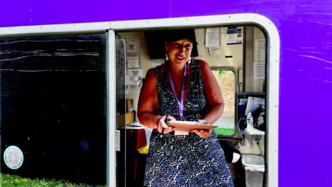 Darren Conway/BBC Karen pictured at work. She's wearing a blue-and-white dot dress with a black baseball cap and purple lanyard. She holds a clipboard and smiles at the camera. 