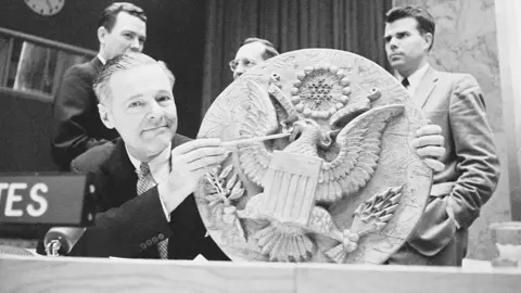 Getty Images A black and white image of US Ambassador to the UN Henry Cabot displaying a wooden replica of the Great Seal of the United States, which Soviet boy scouts presented to US Ambassador Averell Harriman in 1945.