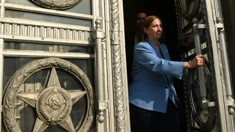 AFP via Getty Images Lynne Tracy walks through a large set of ornate double doors. She has short brown hair and wears a cornflower blue blazer. She is pushing open the left door using a long vertical handle. The doors are engraved with bronze-coloured symbols, including the hammer and sickle.