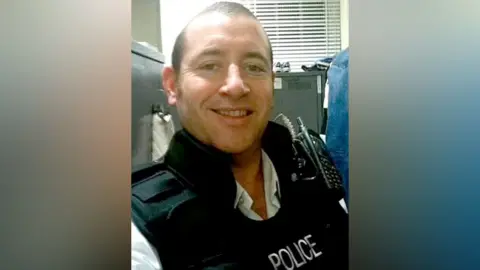 Social media A selfie taken by David Carrick of him sitting in an office with a filing cabinet behind him. He is smiling and has a walkie-talkie strapped to his chest.