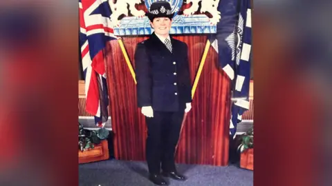 Linda Williams An official photo to mark when Linda xx joined the Metropolitan Police. Linda stands smiling into the camera wearing a full Met Police uniform and white gloves. She is flanked by a Union Jack flag on one side and a Met Police flag on the other. 