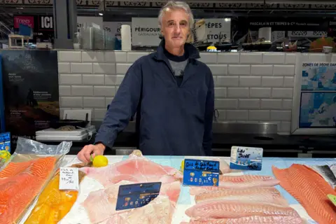 Jean Roger Faucher, a man in his sixties, stands behind his fishmongers stall. Various cuts of fish are in front of him and he wear a navy jacket.