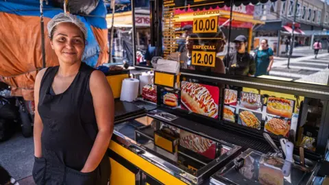 A woman with a hair net stands in front of a mobile hot dog stand
