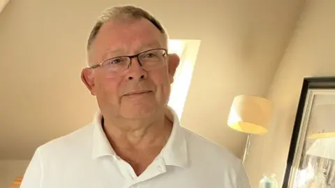 Rod Stone, who is wearing glasses and a white polo shirt, is standing in his living room and is smiling at the camera.