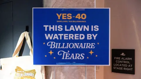 Getty Images A "Yes to 40 This Lawn Is Watered By Billionaire Tears" yard sign among merchandise for sale during a "Yes on 40 for Ballots Over Billionaires" rally in San Francisco, California, US, on Saturday, Oct. 3, 2026. 

