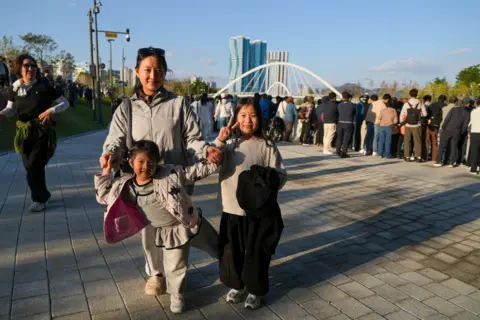 Hosu Lee/BBC A family of three smiling at the camera as a crowd gazes into the canal watching Bukangi. 