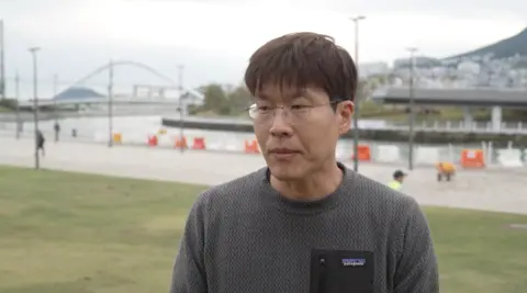 Reuters A person wearing glasses and a grey patterned Patagonia sweatshirt stands on a grassy area beside a waterfront canal in Busan, South Korea. In the background, a pedestrian bridge arches over the water, while construction barriers, lampposts and a riverside walkway line the canal. A hill with buildings is visible in the distance under an overcast sky. Visible text on the clothing reads “Patagonia.”