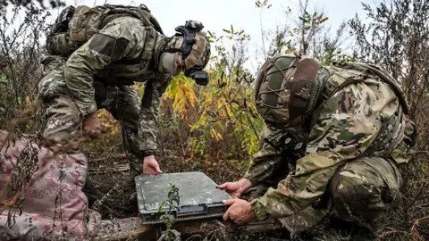 Dmytro Smolienko/ Ukrinform/ Getty Images Two soldiers in green camouflage set up a Starlink terminal, which is also covered in green camouflage.