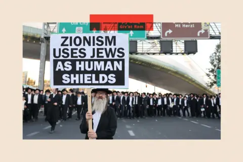 EPA A Haredi man hold a sign saying 'Zionism uses Jews as human shields' under a motorway overpass, with other Haredi men in the background