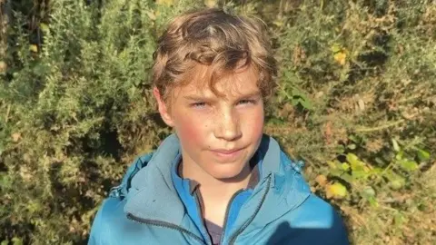 Liam has blonde curly hair and is smiling. 
He is wearing a blue jacket and blue fleece. 