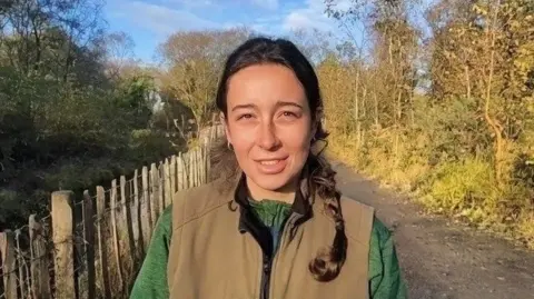 Chloe McElhatton has long brown hair and is smiling. She is wearing a tan gillet and green shirt. She is standing on a path with a wooden fence and large trees.