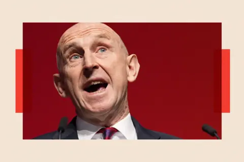 PA Media John Healey wearing a white shirt with a red and purple tie, speaking at a podium into microphones.