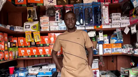 Nabila Muftawu A man wearing a beige kaftan matching set, standing inside an electrical supplies shop, hands in his pockets, smiling into the camera