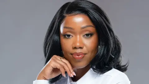 Cris Njoki A studio head shot of a woman wearing full-face makeup and a bob wig, wearing a white shirt, looking directly into the camera while resting her head on the back of her right hand