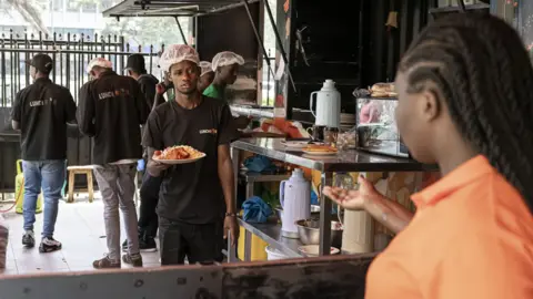 Bloomberg via Getty Images A man wearing a black T-shirt, black trousers and a white hairnet stands behind a food counter, handing a plate of food to a female waiter in an orange polo shirt.