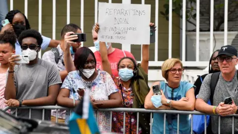 Toby Melville - Pool / Getty Images  A woman holds a sign in protest as she waits for the arrival of Britain's Prince William and Catherine, Duchess of Cambridge, on the seventh day of their tour of the Caribbean on March 25, 2022 in Nassau, Bahamas