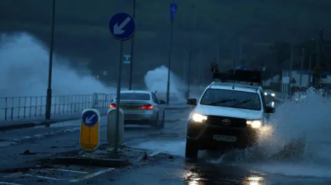 Reuters Cars driving along a coastal road which has large puddles. The picture is dark suggesting it was taken at night time.