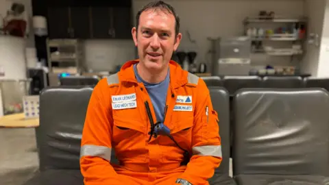 Ewan Leonard is sitting down on a grey seat. He has dark, receding hair and is wearing an orange boiler suit with his name on it. He is smiling.