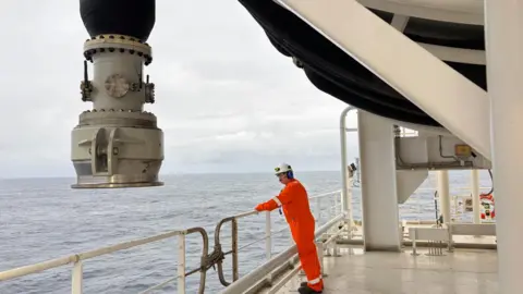 Man in orange overalls and a white helmet leans over a railing with the sea beyond. There is a large nozzle hanging down near him