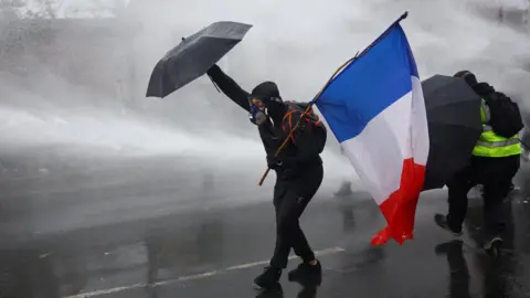Reuters A masked prostester holding an umbrella and a French flag reacts as French police use a water canon amid clashes during an authorised demonstration organised by labour unions in Paris.