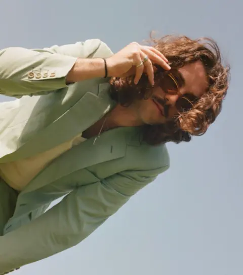 J Casa J Casa - a man with long brown hair and a moustache touches his sunglasses while bending down towards the camera. He is wearing a light green uit and a light yellow v neck top.