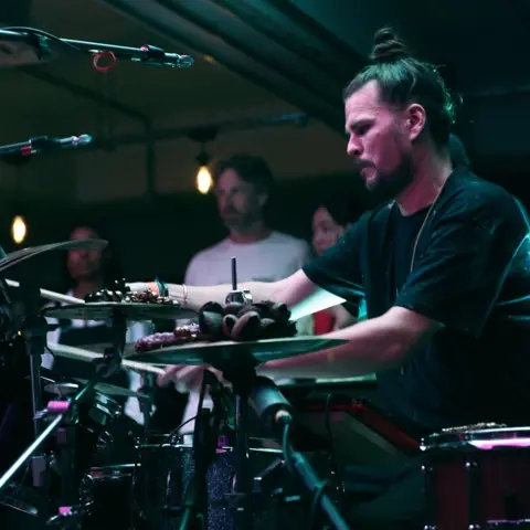 Jordan Matyka A man with a hair bun sitting at a drum set. He is wearing a black t-shirt and on a stage with purple and blue lighting.