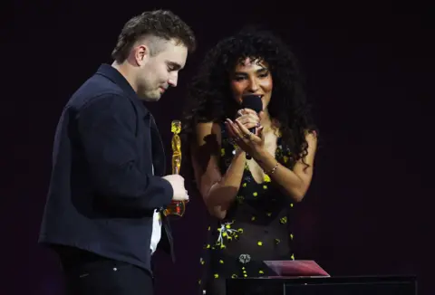 Reuters Olivia Dean speaks on stage next to Sam Fender, during the BRIT Awards at the Co-op Live Arena, in Manchester, Britain, February 28, 2026. 