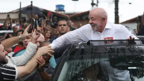 EPA/ Shutterstock Lula, with a white beard and in a white button-down shirt, smiles as he stands through the sunroof of a rain-speckled vehicle, reaching out to shake hands with supporters who are stretching their arms and holding up smartphones to record the moment. The scene takes place on a crowded street. Behind him a concerned looking man watches closely (a security agent?)
