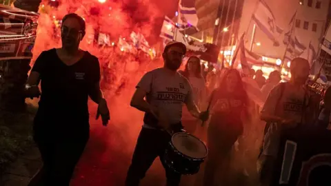 AFP via Getty Images Protesters bang a drum and light red flares. It is night. 