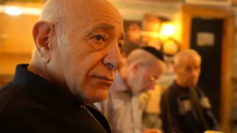 Shlomi sits with friends and looks toward the camera. He is wearing a black jacket. 
