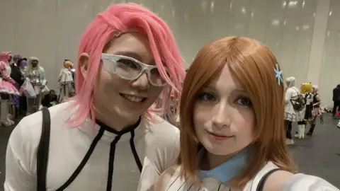 Supplied Nicholas on the right wears a white bodysuit with black piping and a pink wig. Ruby wears a blue collar and white striped outfit and a brown wig. They both look at the camera and smile.