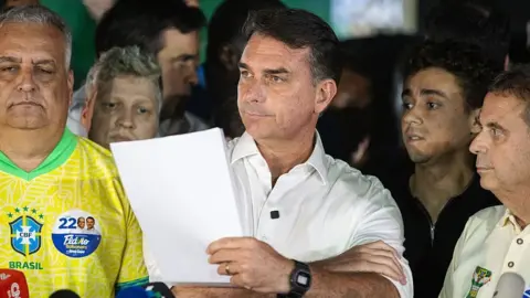 Bloomberg via Getty Images Bolsonaro standing while holding a wad of paper. He is surrounded by people who do not look happy
