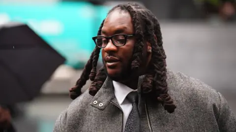 Press Association Omololu Akinlolu arriving at Southwark Crown Court, south London on Monday October 26, 2026. He has shoulder-length hair, glasses and is wearing a grey herringbone jacket, white shirt and grey tie.