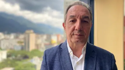 Political analyst Beningno Alarcon, a middle-aged man, is wearing a blue suit over a white shirt. The blurred backdrop of a city's skyline is also in view.