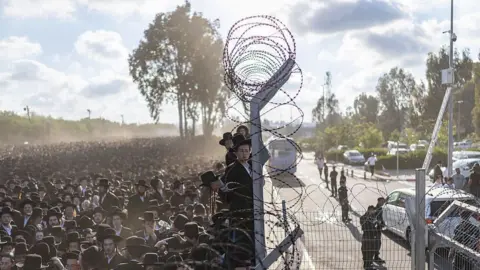 AFP via Getty Images Hundreds or thousands of ultra-Orthodox jews stand beside barbed wire fencing. 
