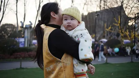 Getty Images A woman in a golden vest holds a baby wrapped in a polka-dot blanket in the park at dusk