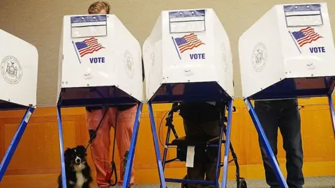 Getty Images Three people voting behind voting booths in New York with a dog sitting at the feet of the person on the left.