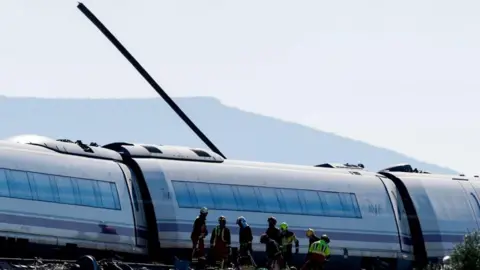 EPA A piece of rail is seen emerging through the roof of a train carriage; there are emergency workers wearing high-visibility jackets and helmets in front of that carriage - one of three visible in the shot.