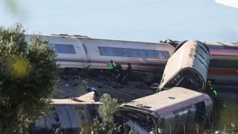 EPA Three overturned train carriages lying on an embankment, with others from another train tilted over. Emergency workers can be seen helping people