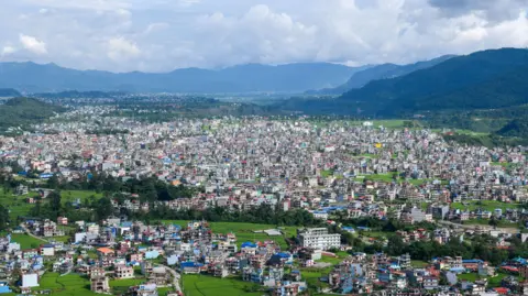 Piyas Biswas/SOPA Images/LightRocket via Getty Images A panoramic view of Pokhara, Nepal, showing a dense spread of homes and low-rise buildings interspersed with bright green fields across a broad valley. Forested hills and mountains surround the city in the distance beneath towering clouds and a hazy blue sky.